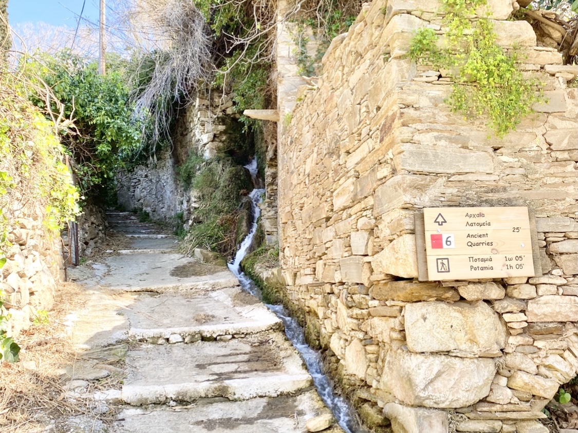 A wide path runs up the side of a stone ruin with a wooden waymarker sign and a spring running next to it | Hiking in Naxos Greece | greekislandbucketlist.com